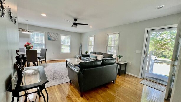 A spacious living room with hardwood flooring, a black leather sofa, a dining area with a glass table, and a ceiling fan.