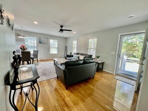 A spacious living room with hardwood flooring, a black leather sofa, a dining area with a glass table, and a ceiling fan.