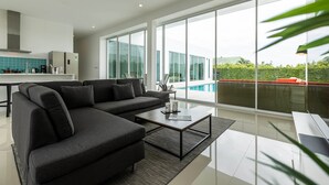 A modern living room with a large sectional sofa, a coffee table, and a view of a swimming pool through large windows. - Modern 4 Bedroom Pool Villa - KHA5 (Hua Hin)