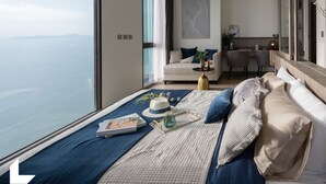 Une chambre à coucher moderne avec une grande fenêtre, un lit recouvert d’une couverture bleue et blanche, une table de chevet avec un livre et un chapeau, et une vue sur la mer.
