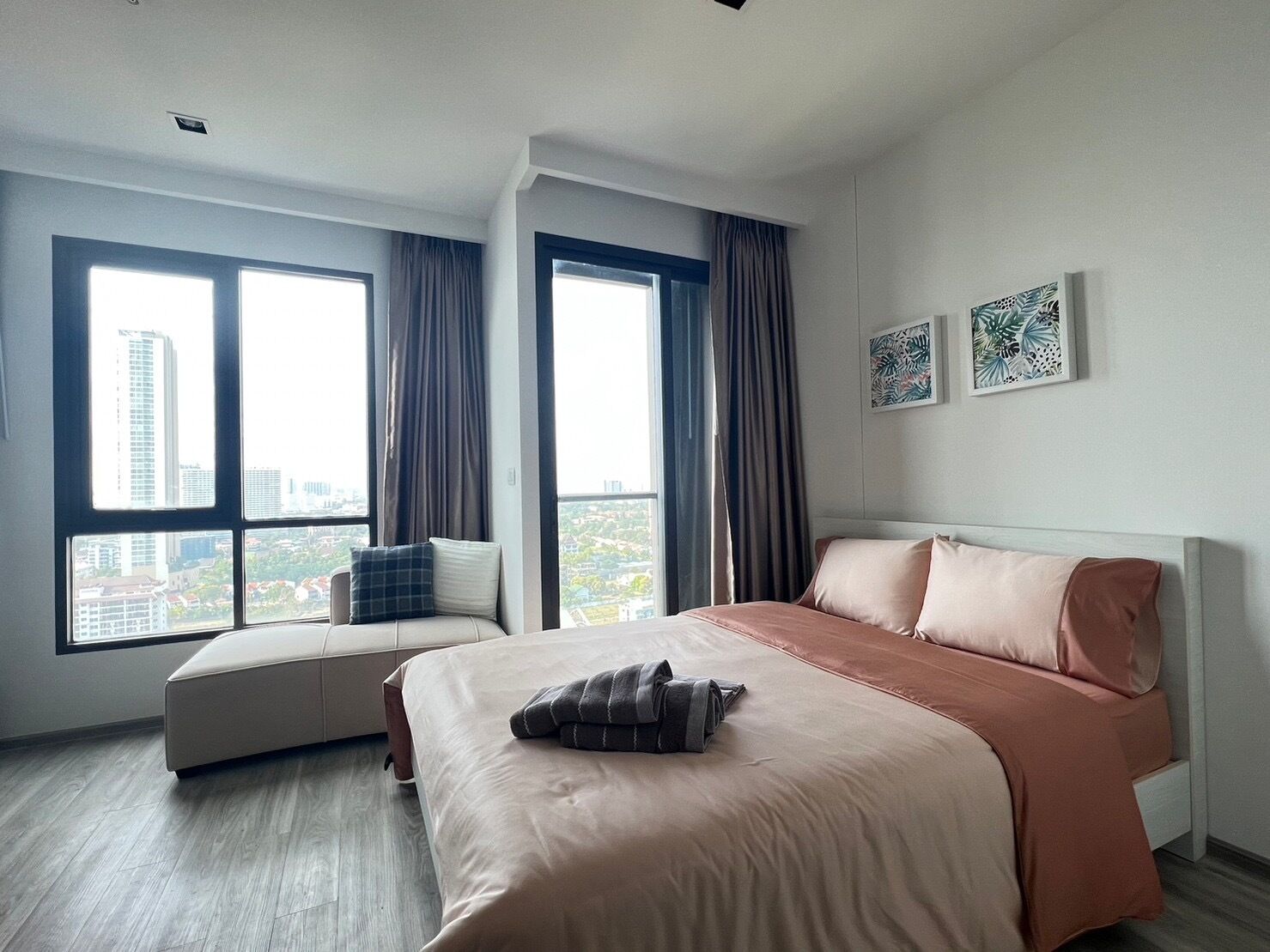 A modern hotel room with a large bed, a sofa, and two windows offering a city view.