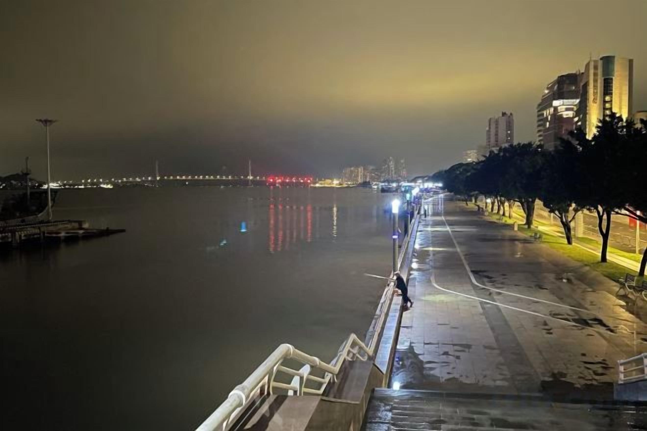 Eine Uferpromenade mit Geländer, ruhigem Gewässer und in der Ferne Stadtlichtern.
