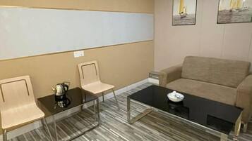 A modern living room with a beige sofa, a glass coffee table, and two chairs. There are framed pictures on the wall.