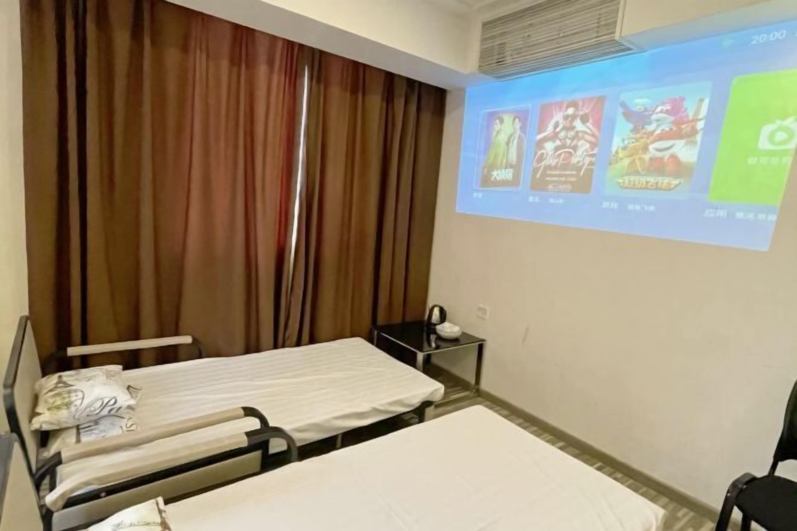 A hotel room with two beds, a small bedside table, a projector screen displaying movie posters, and a window with curtains.