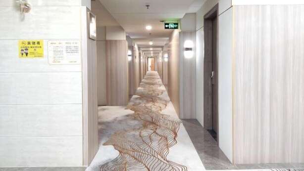 A long, narrow hallway with a patterned carpet, doors on both sides, and a sign on the left wall. - Fulin Hotel (Huilai)