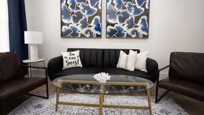 Un salón con un sofá de cuero negro, dos cuadros abstractos en la pared, una mesa de centro de vidrio con un centro floral y una alfombra estampada.