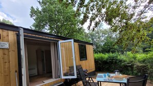 Una casa móvil de madera con la puerta abierta que muestra una habitación con una cama y un ventanal. En el exterior, hay una terraza con una mesa, sillas y una cafetera.