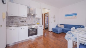 Una cucina moderna con mobili bianchi, forno integrato e schienale in piastrelle. C'è una zona pranzo con tavolo e sedie, e un divano blu nel soggiorno.