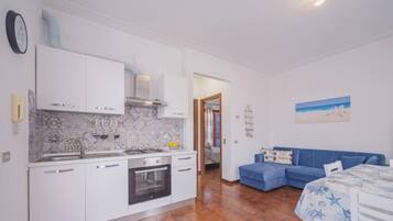 A modern kitchen with white cabinets, a built-in oven, and a tiled backsplash. There is a dining area with a table and chairs, and a blue sofa in the living room.