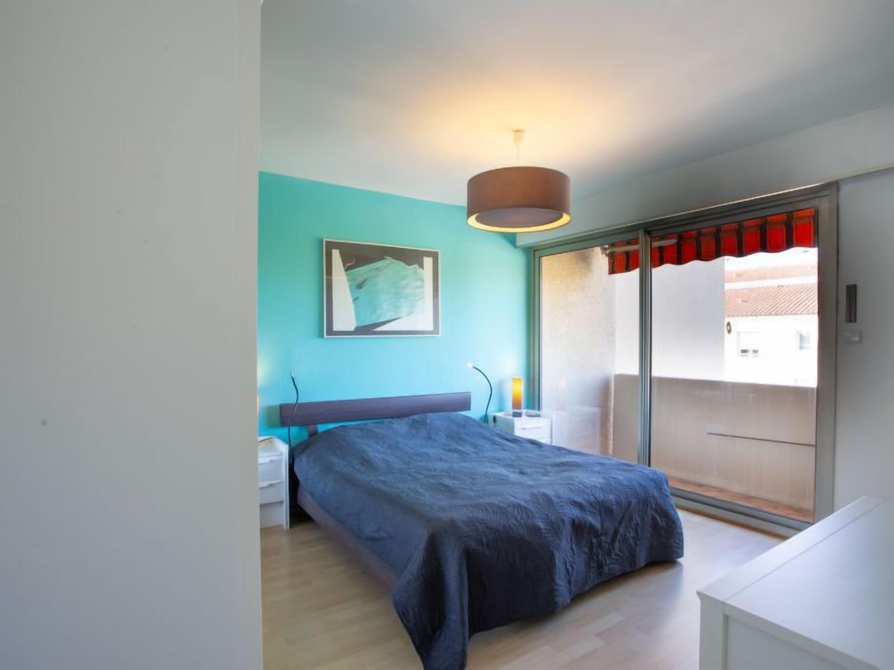 Un dormitorio con cama, mesita de noche, lámpara, un cuadro en la pared y un ventanal corredero que da a un balcón.