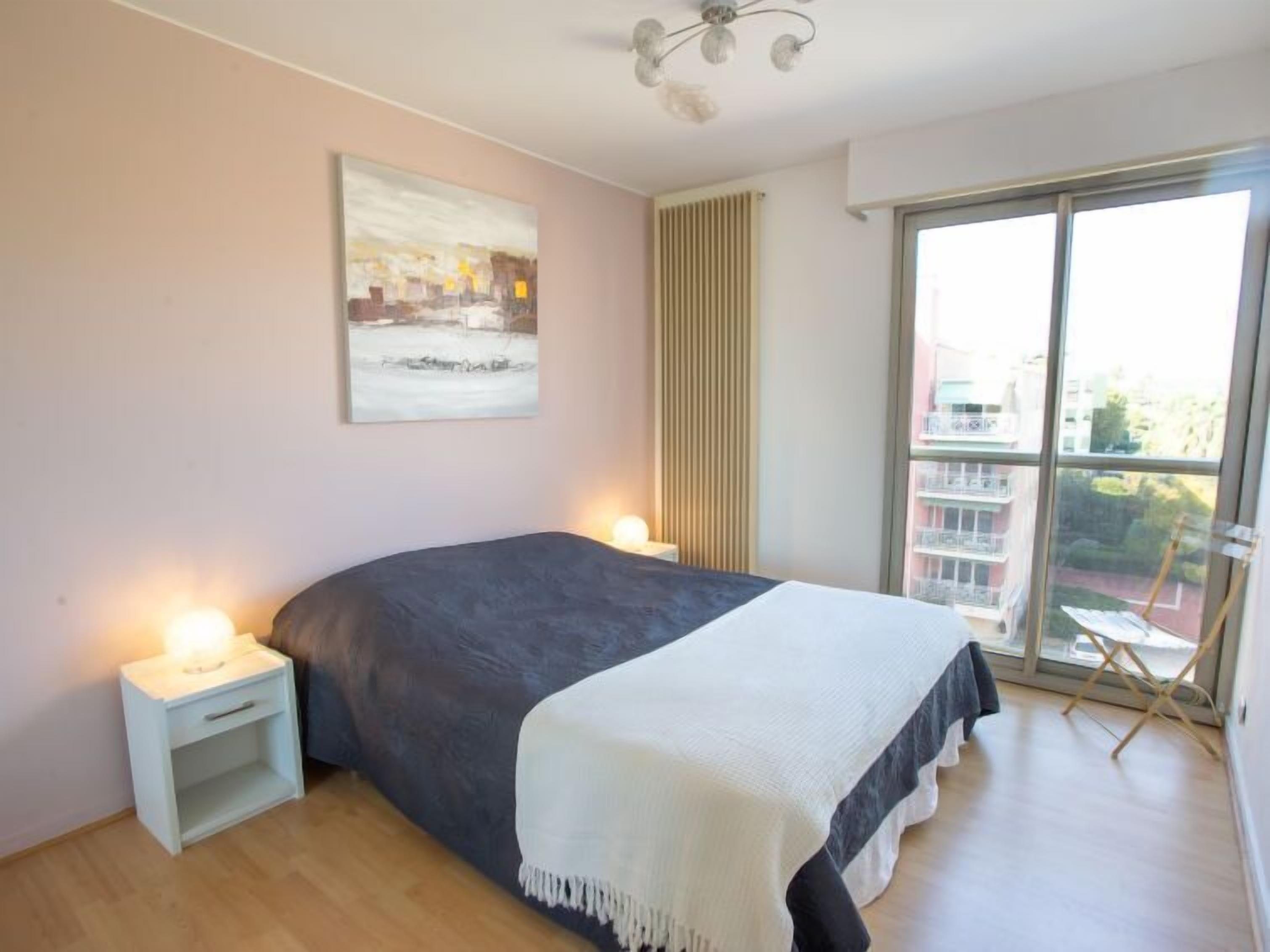 A bedroom with a bed, bedside tables, a painting, a window with a view, and a ceiling light.