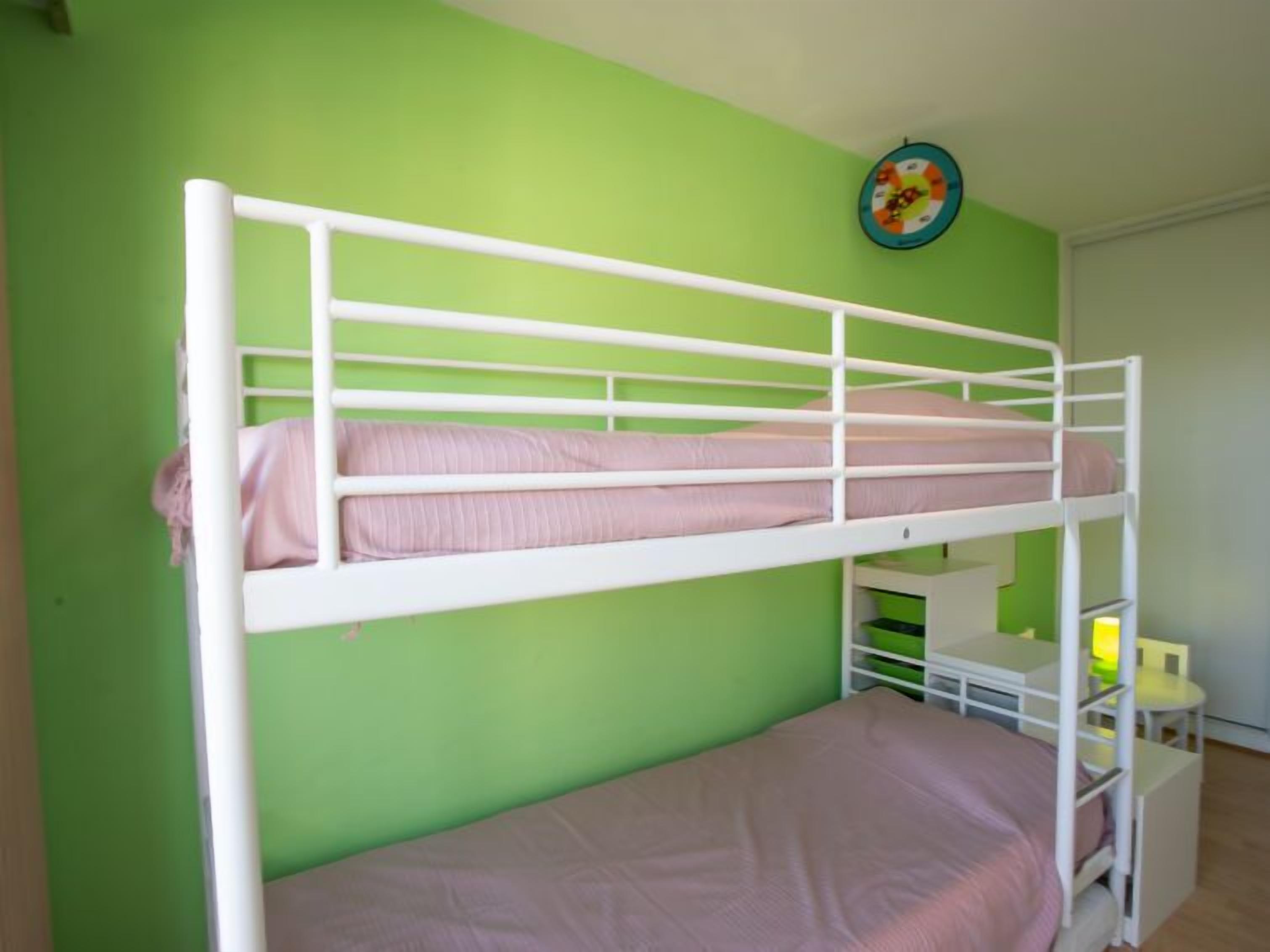 A bunk bed with a lower bunk and a single bed, green walls, a small table with a lamp, and a clock on the wall.