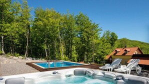 A hot tub surrounded by lounge chairs and a swimming pool, set against a backdrop of trees and a hillside.