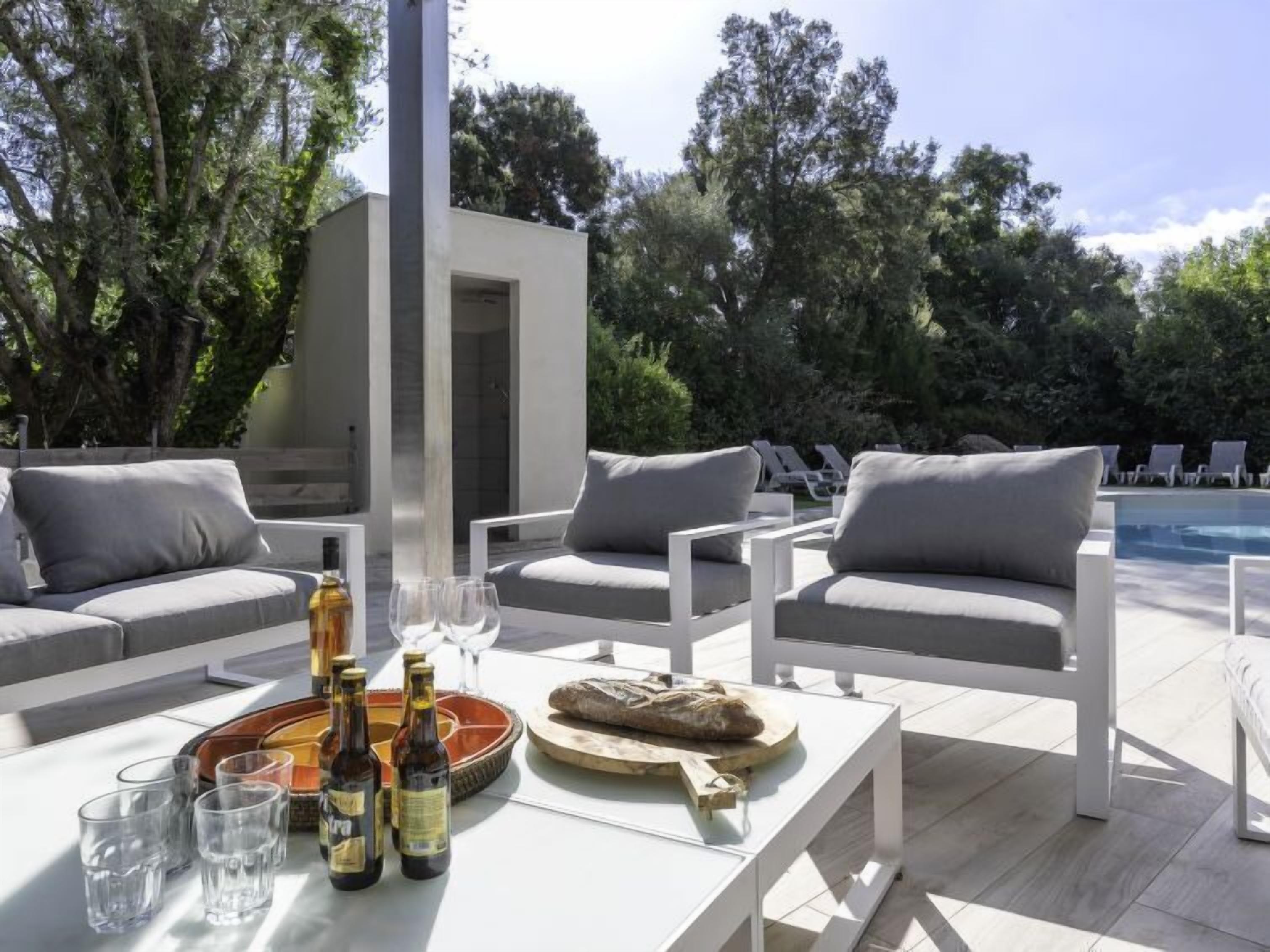 Un área de estar al aire libre con mobiliario blanco, una piscina y una pequeña construcción.