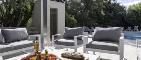 Un área de estar al aire libre con mobiliario blanco, una piscina y una pequeña construcción.