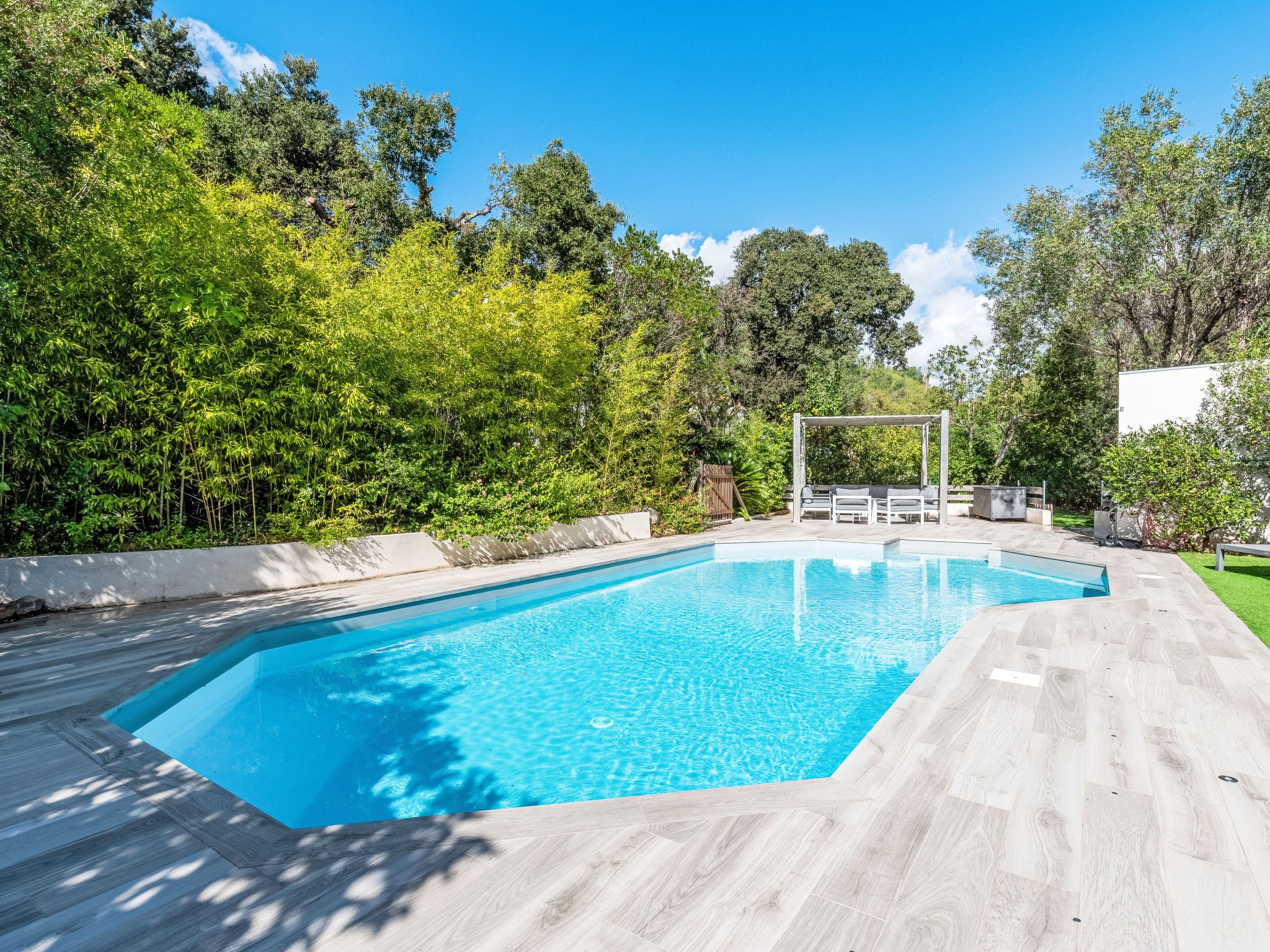 Una piscina rectangular con una cubierta de madera que la rodea, frente a un fondo de vegetación exuberante y árboles.