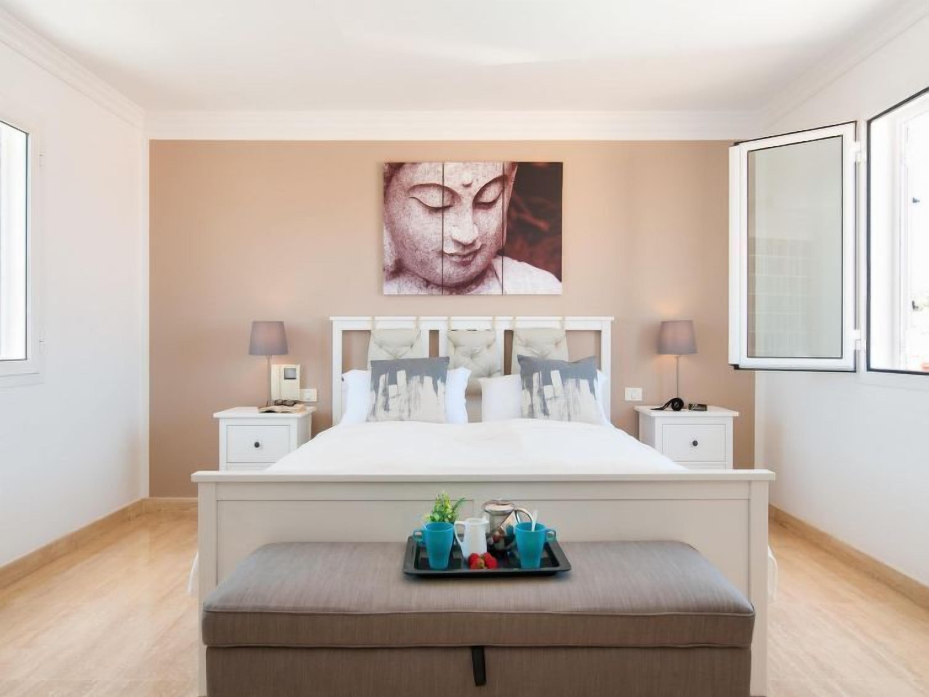 A bedroom with a large bed, two bedside tables, a bench with a tray of fruits and a plant, and a large wall art piece.