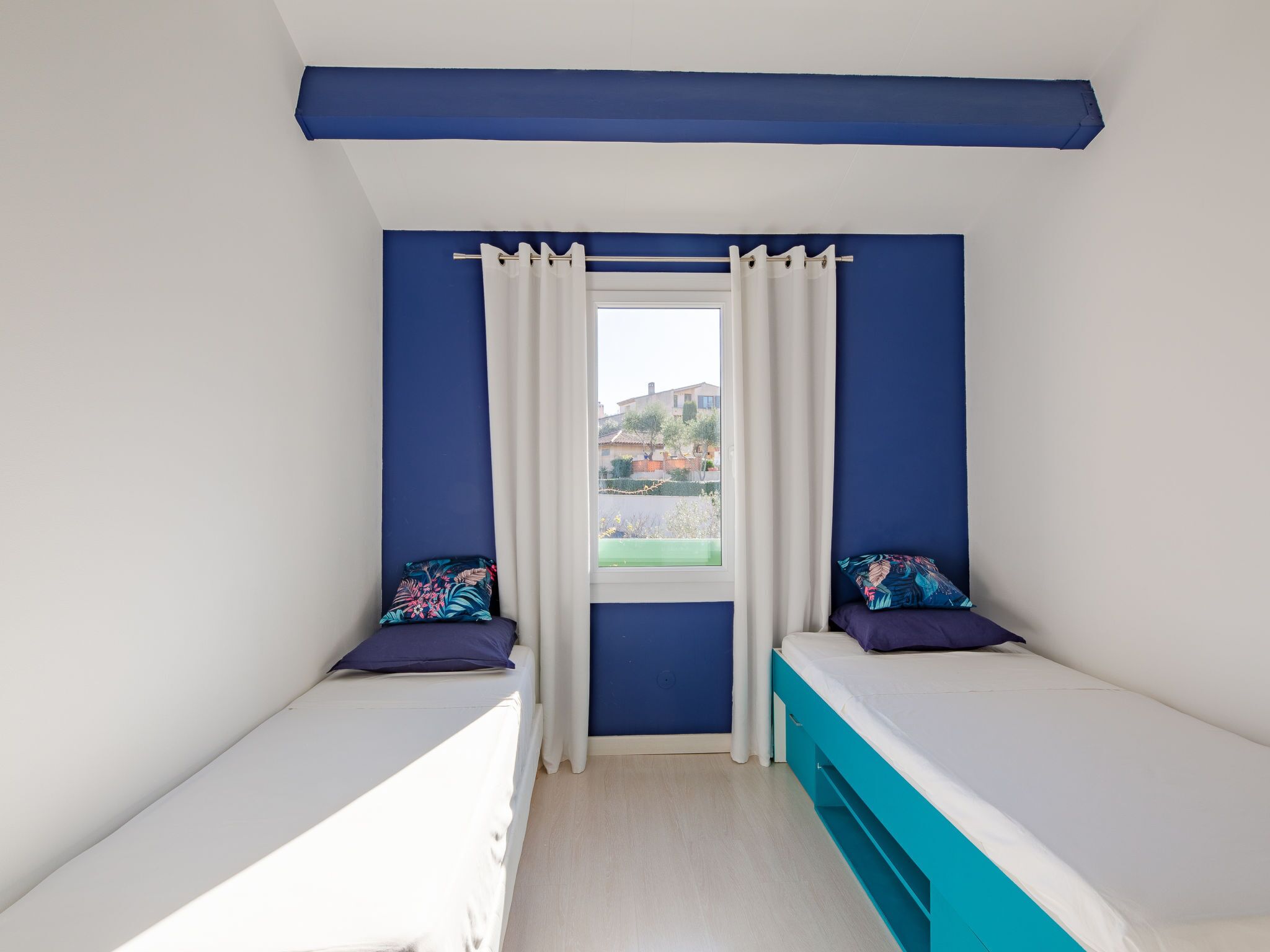 A room with two beds, a window with curtains, and a view of the outside.