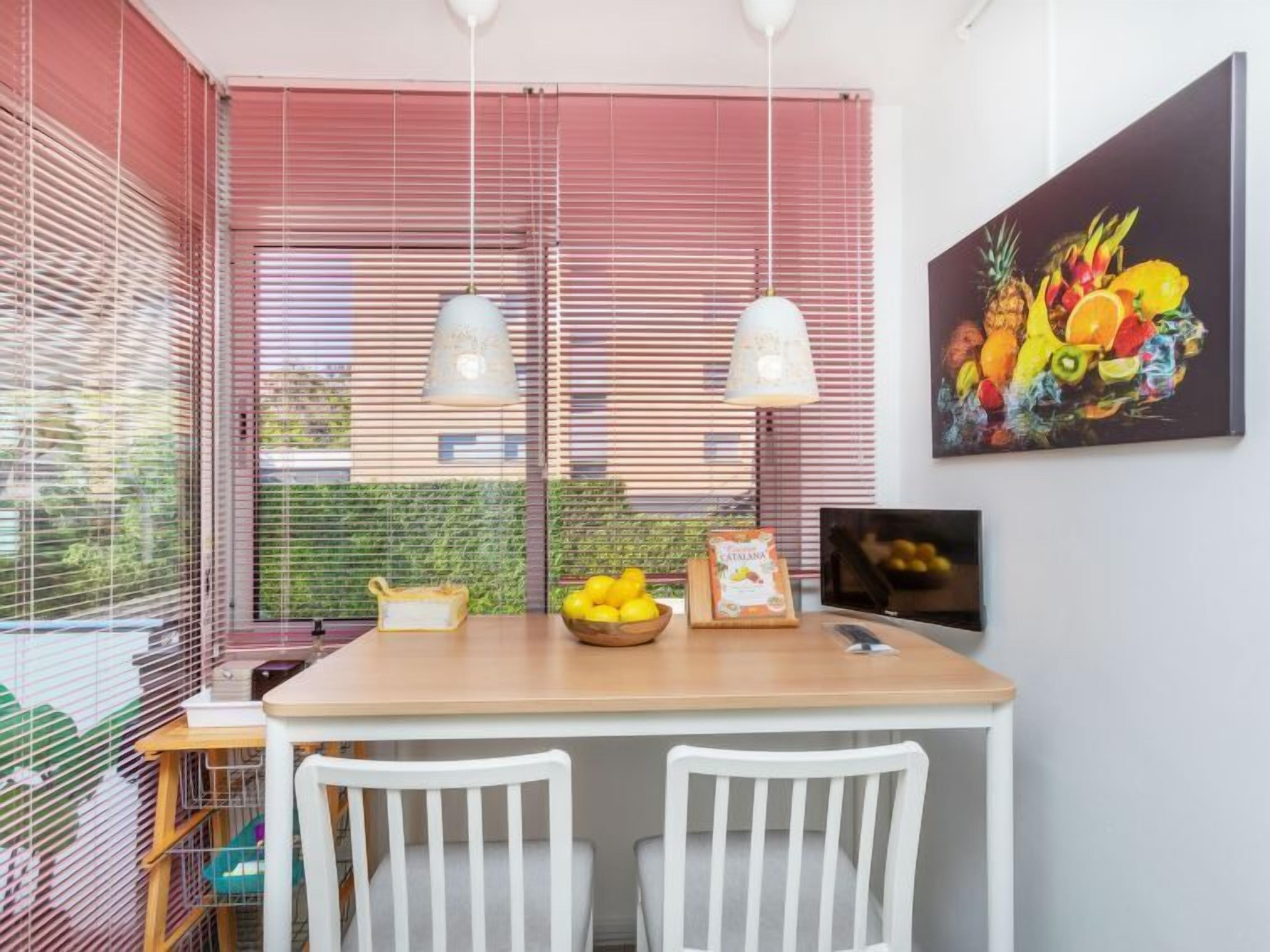 A dining area with a wooden table, white chairs, a fruit bowl, and a painting of fruits on the wall.