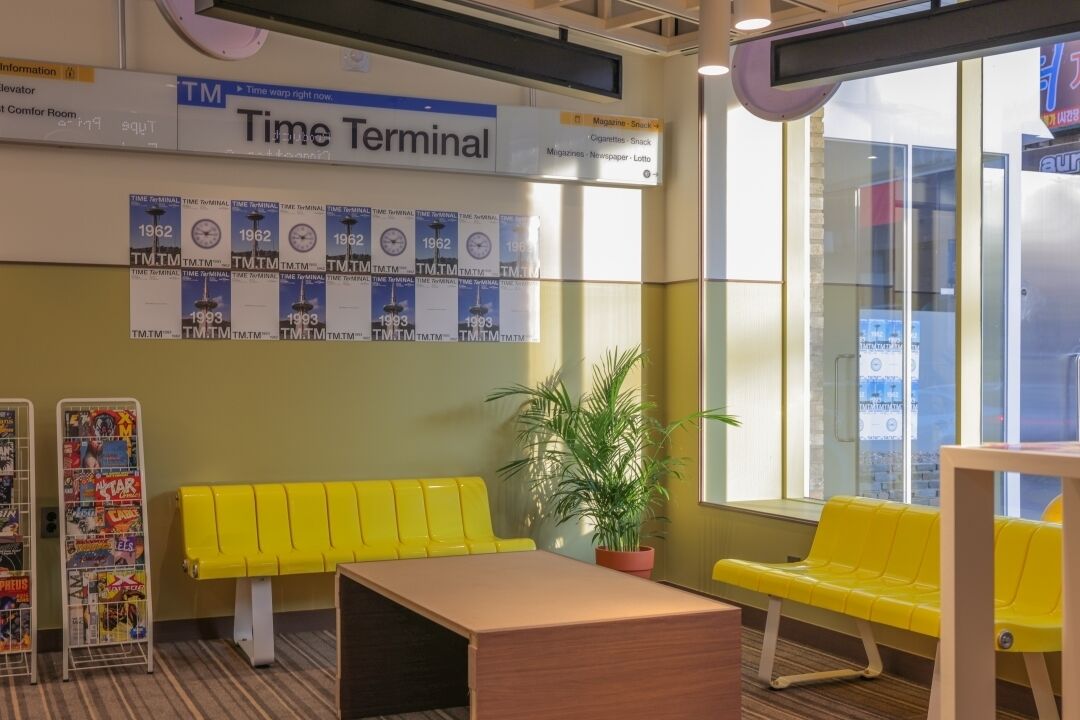 A Time Terminal with a yellow bench, a potted plant, and a wall displaying Time Terminal posters from 1962 and 1993.