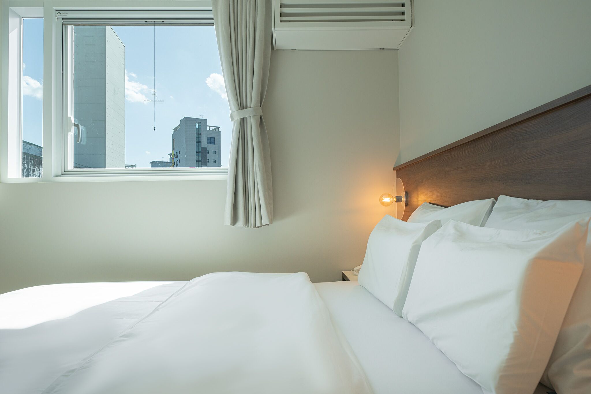 Ein Hotelzimmer mit einem großen Bett, einem Fenster mit Vorhängen, einem Nachttisch mit Lampe und Blick auf umliegende Gebäude.