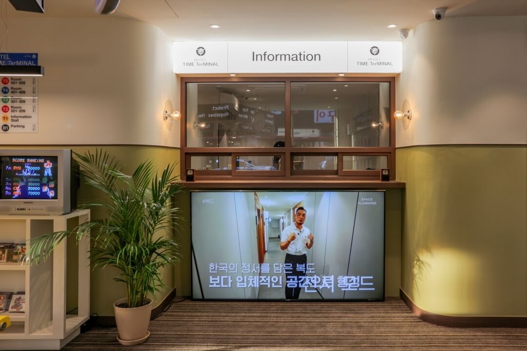 A hotel reception area with an information desk, a television screen displaying a program, and a potted plant.