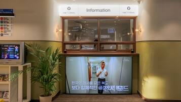 A hotel reception area with an information desk, a television screen displaying a program, and a potted plant.