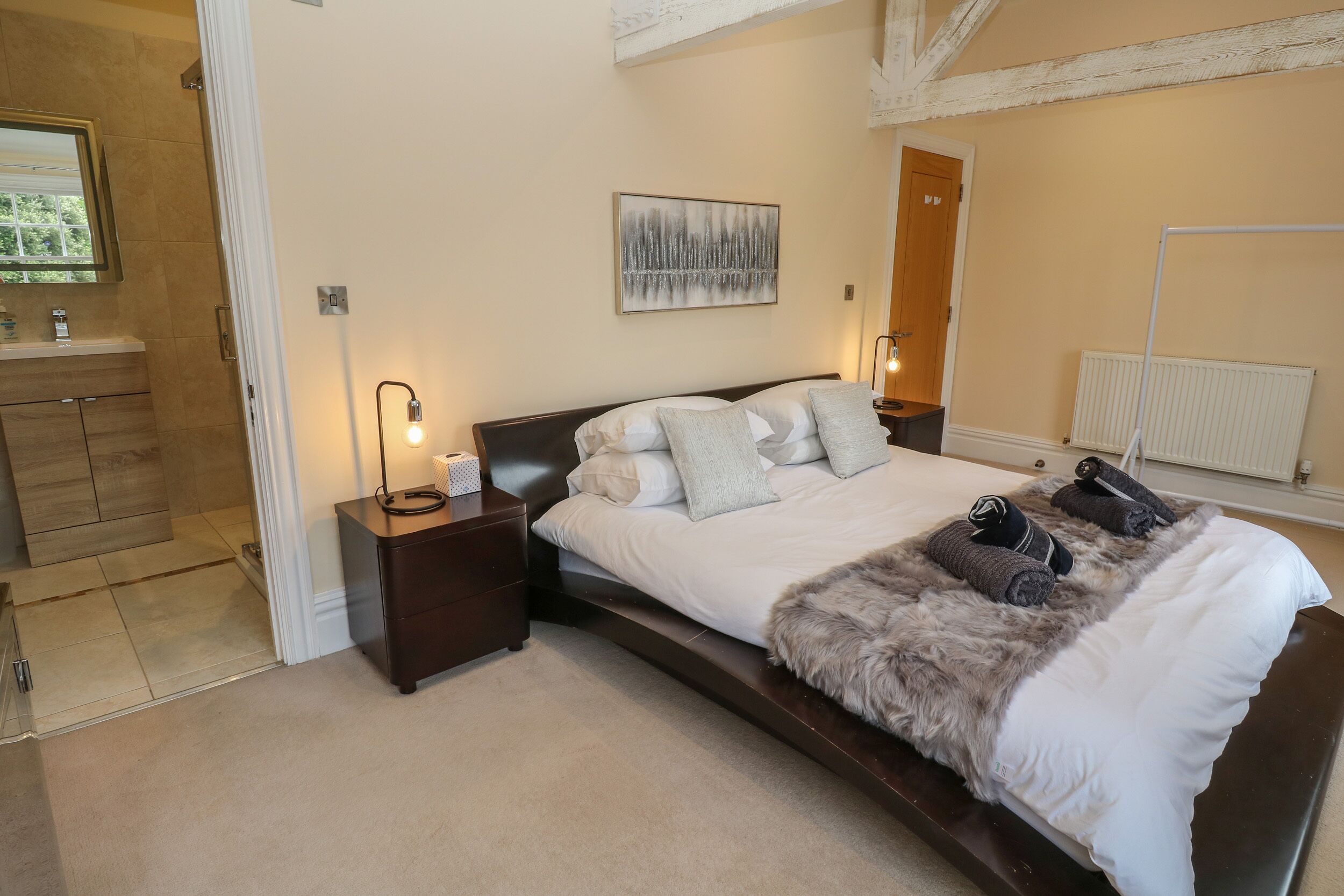 A bedroom with a large bed, two bedside tables, a nightstand, a fur throw, and a painting on the wall.