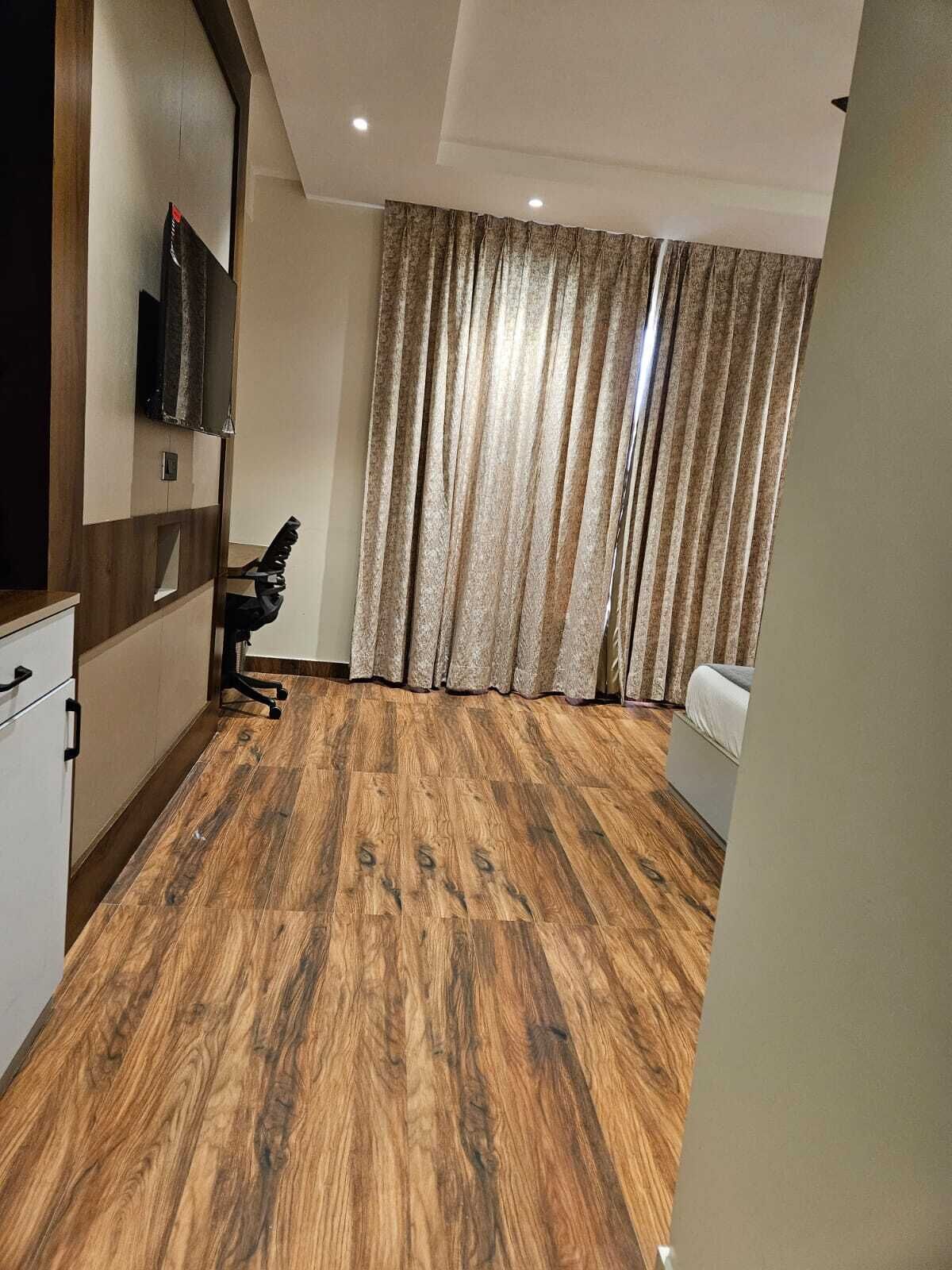 A room with a wooden floor, a bed, a desk with a chair, a television mounted on the wall, and a window with curtains.