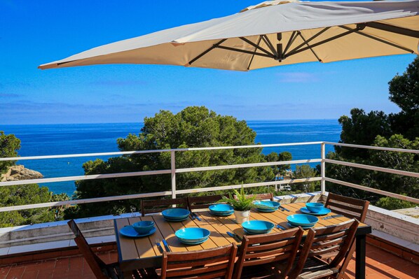 Mesa posta com pratos azuis e um guarda-sol bege em terraço com vista para o mar.