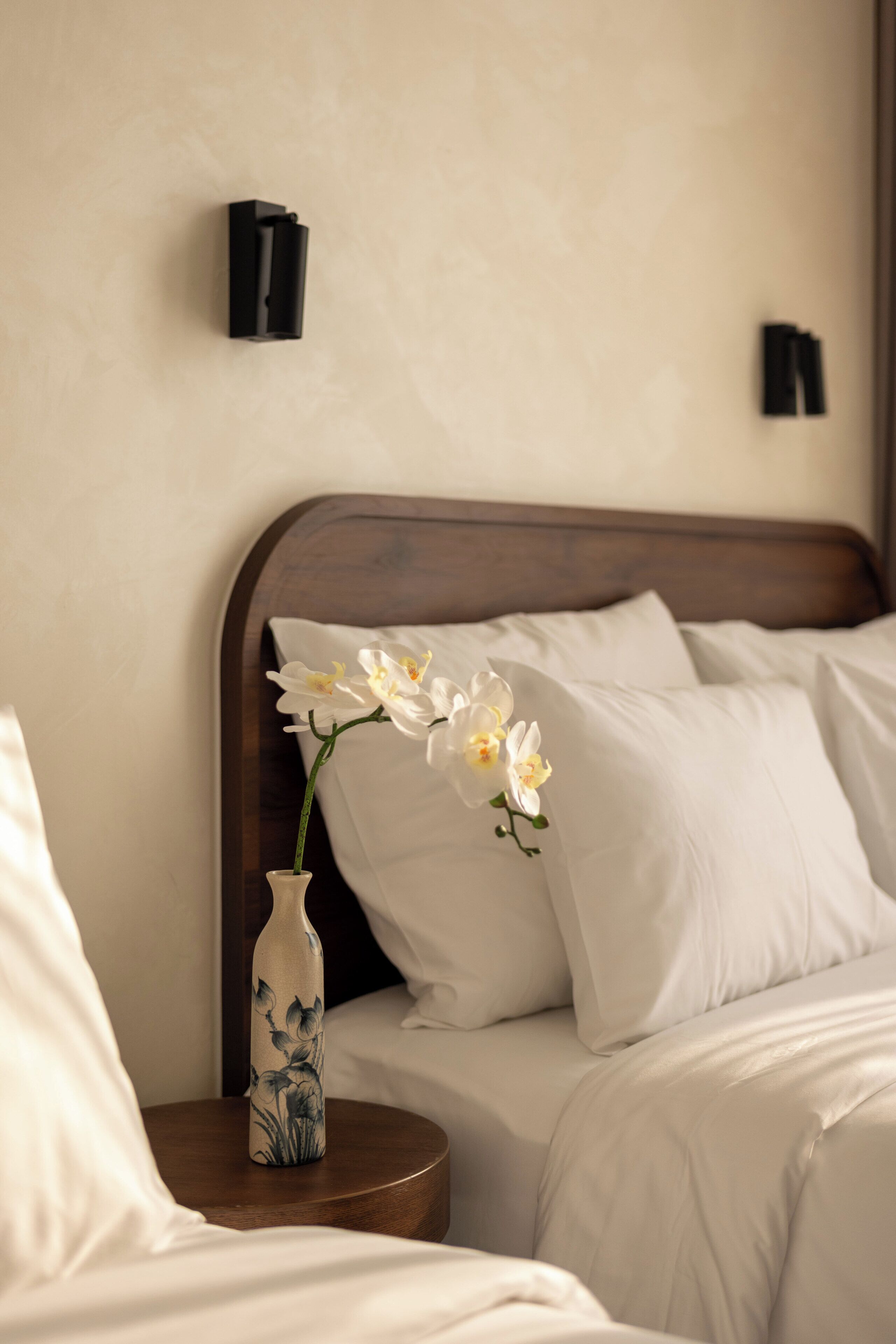 Una cama bien tendida con sábanas blancas, un florero con flores blancas sobre una mesita de noche y dos lámparas de pared.