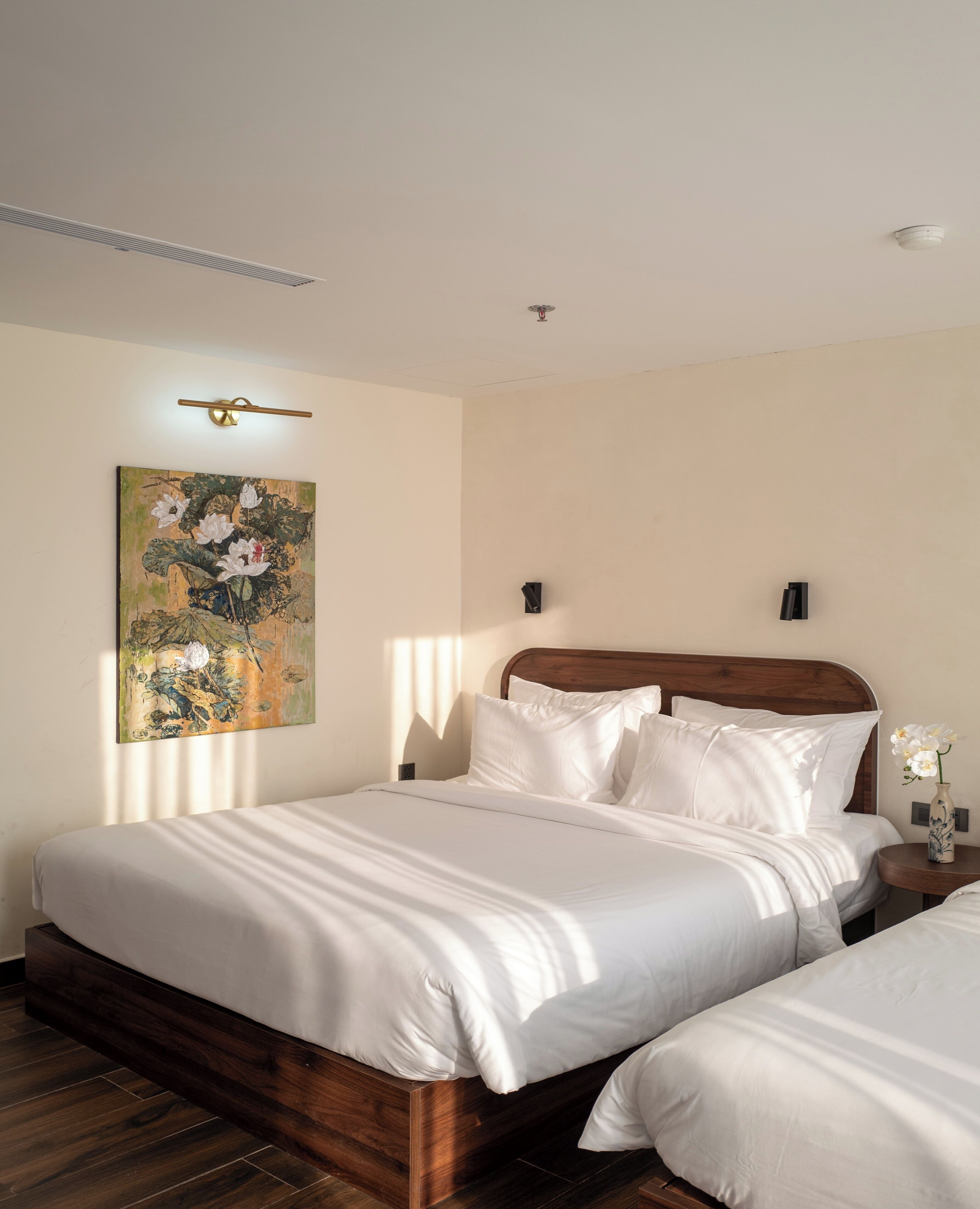 A hotel room with a wooden bed, white bedding, a painting on the wall, and a small table with a vase.