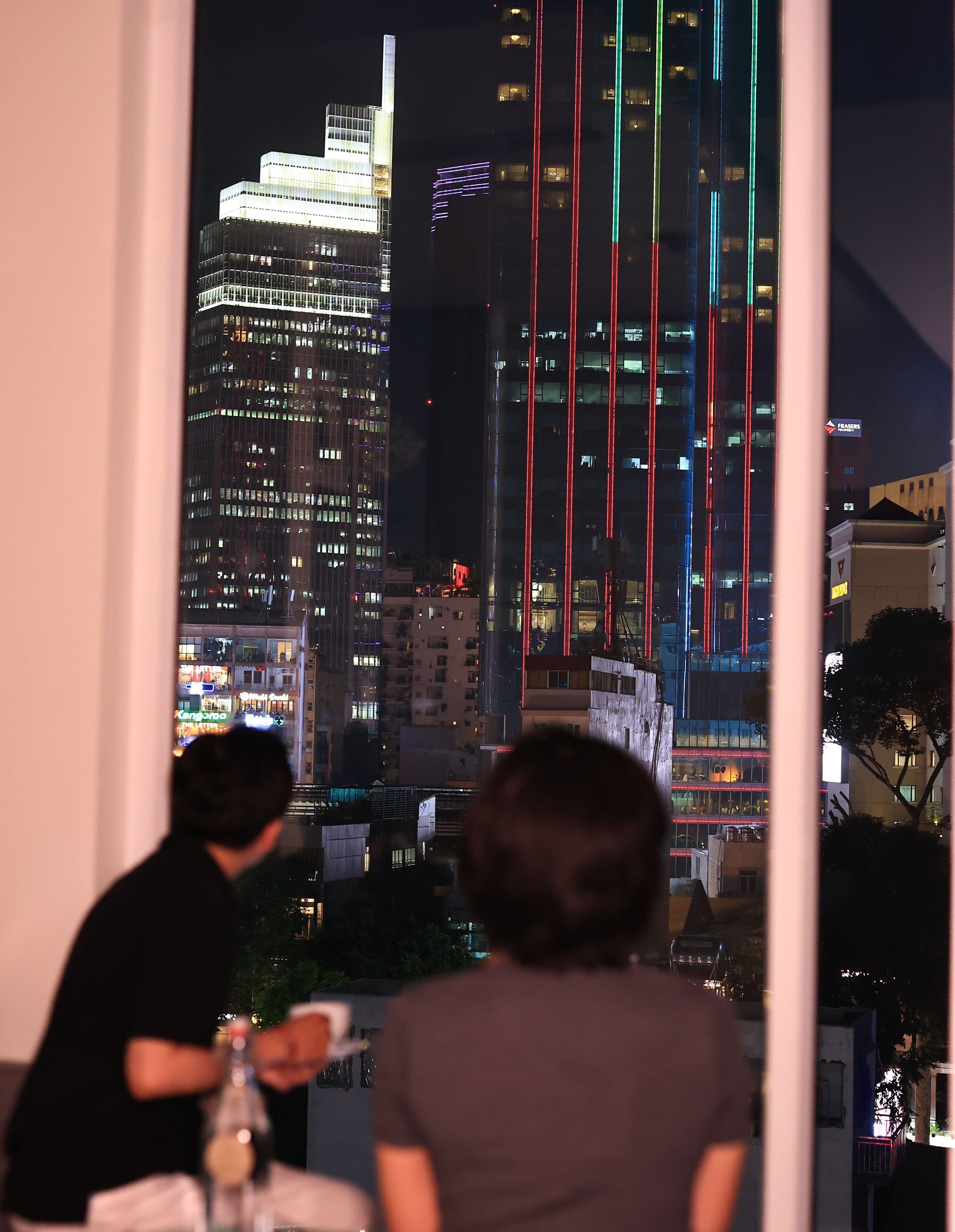 A view from a window overlooking a cityscape at night with illuminated buildings and a person seated at a table.