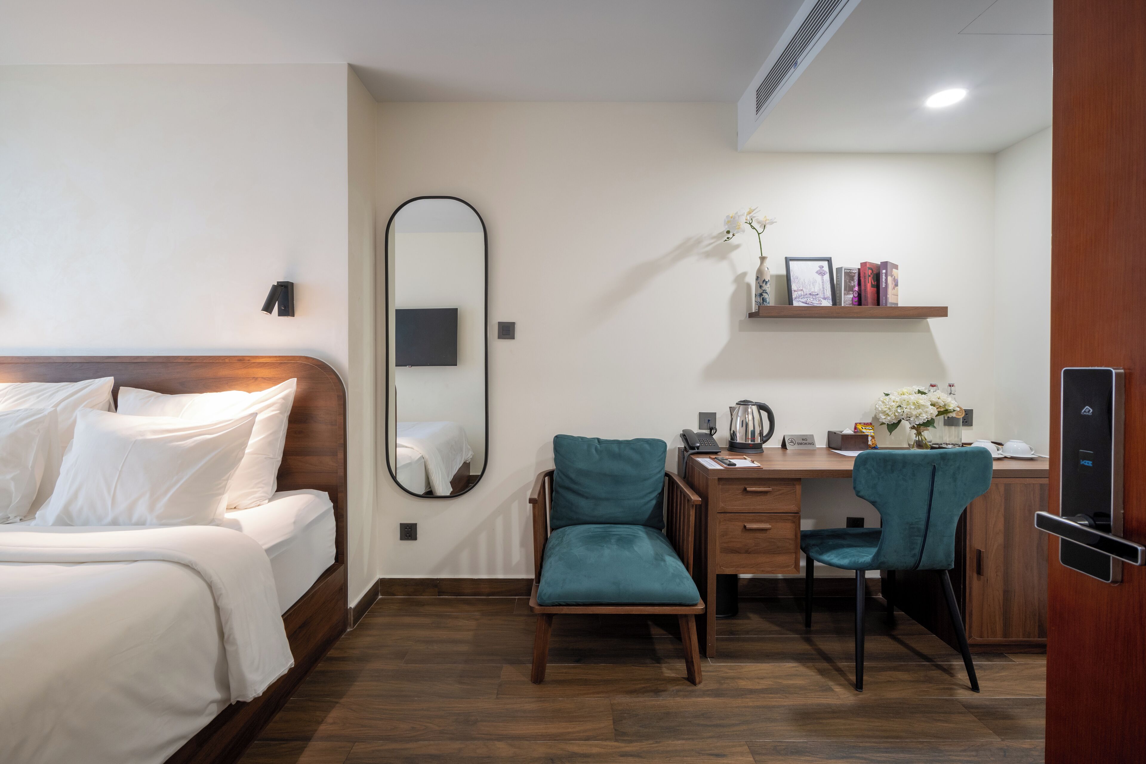 Una habitación de hotel con una cama, un escritorio con dos sillas, un espejo y un estante con libros y flores.