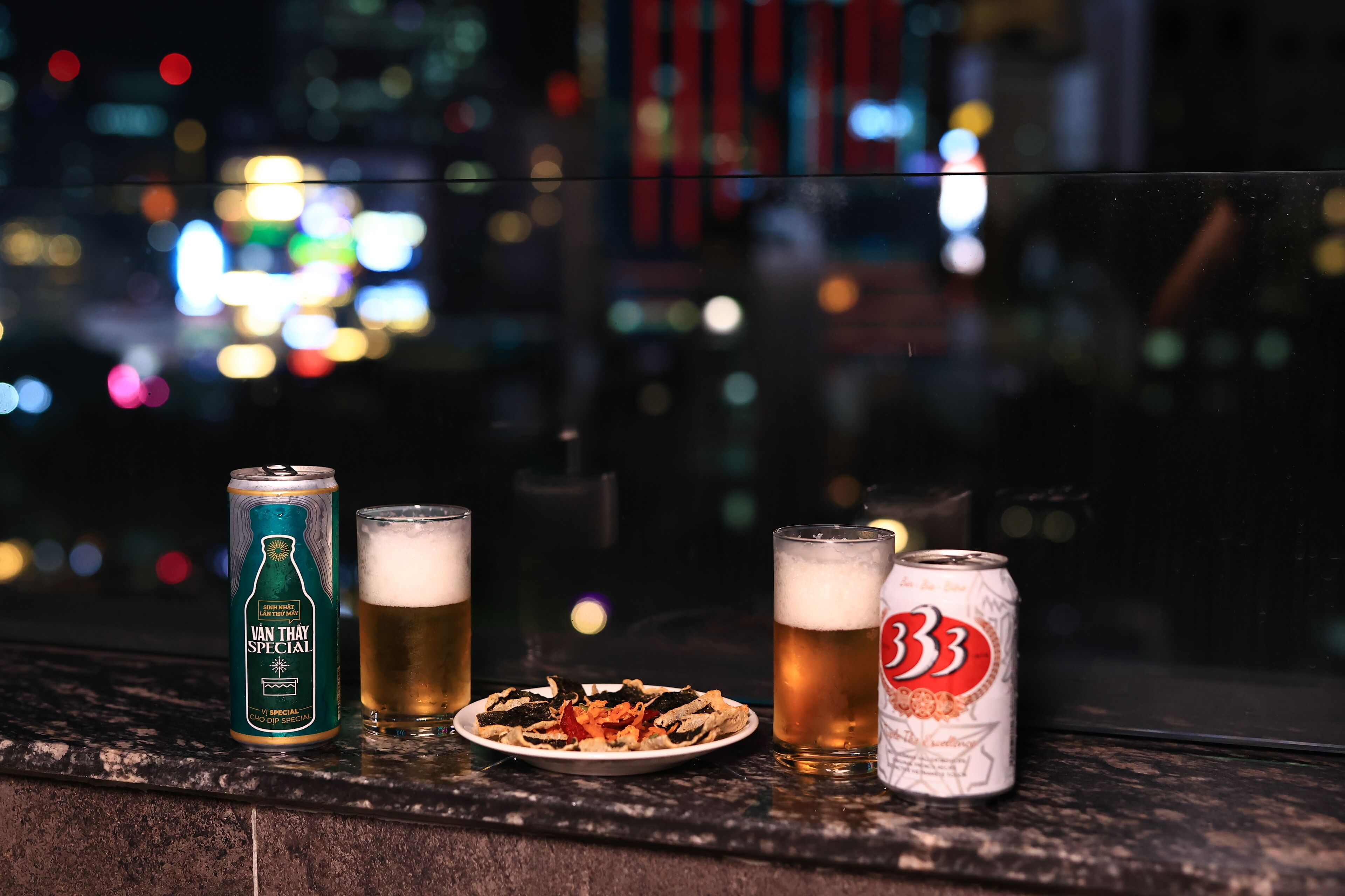 A can of Van Thay Special beer, two glasses of beer, and a plate of food on a window sill with a cityscape view.