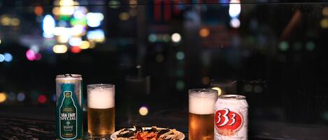 A can of Van Thay Special beer, two glasses of beer, and a plate of food on a window sill with a cityscape view.