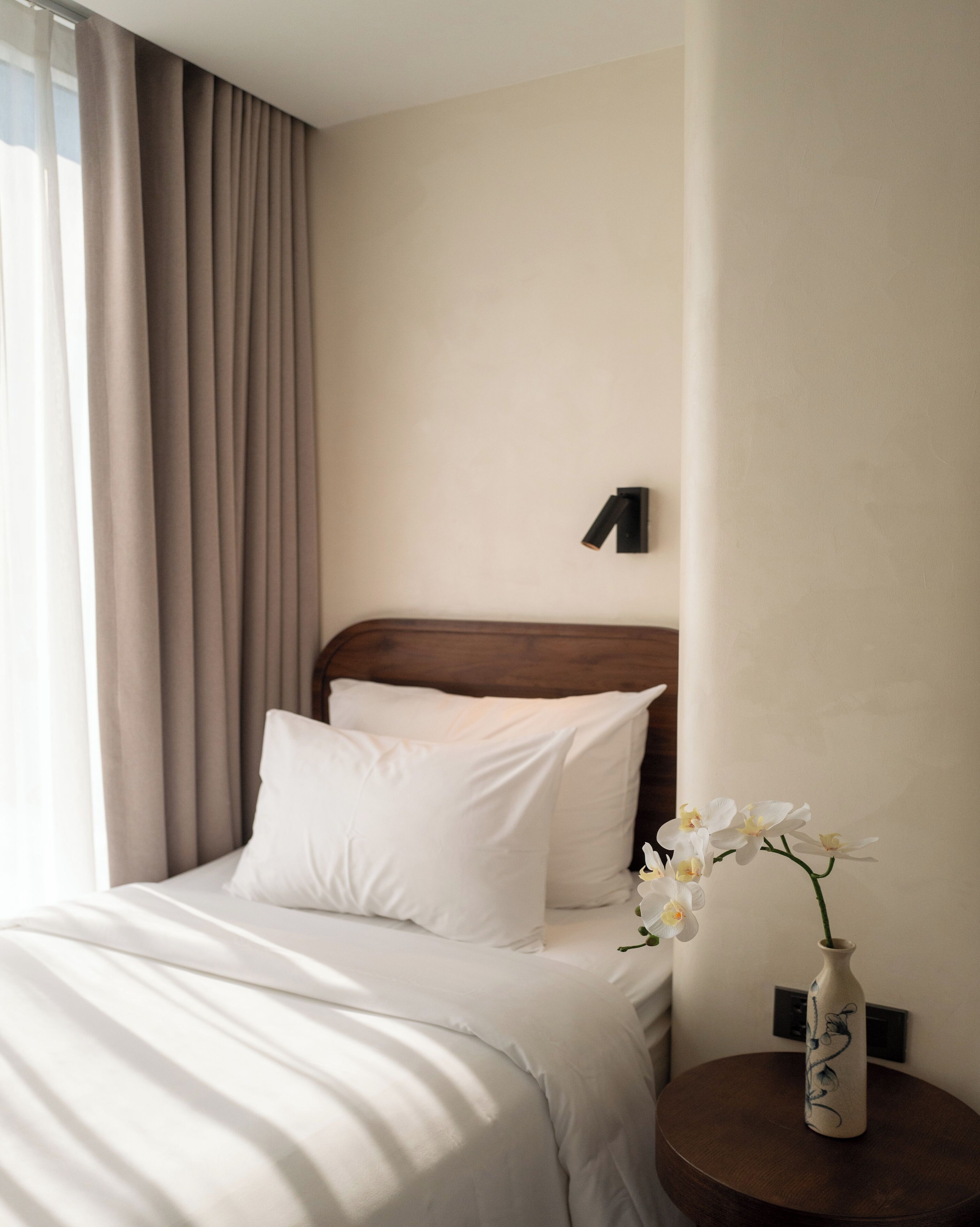 A neatly made bed with white linens, a wooden headboard, and a vase with white flowers on a small round table beside it.