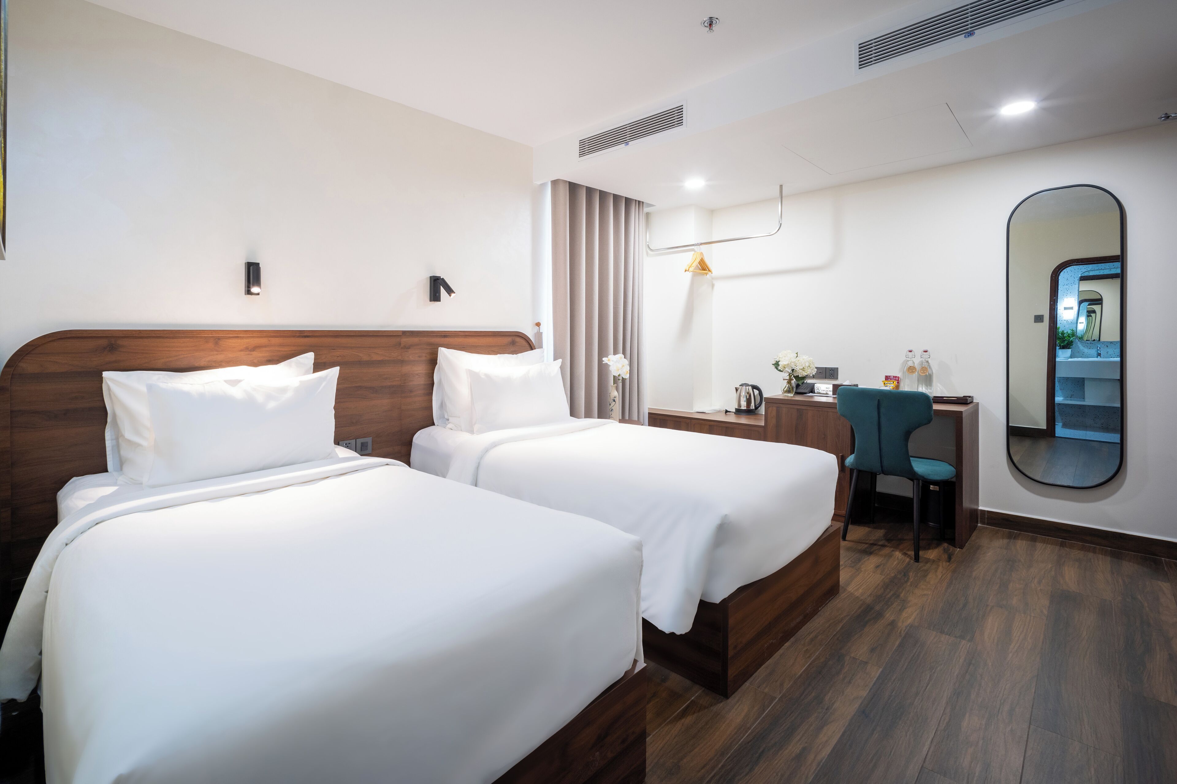 A hotel room with two beds, a desk, a chair, a mirror, and a bathroom visible in the reflection.