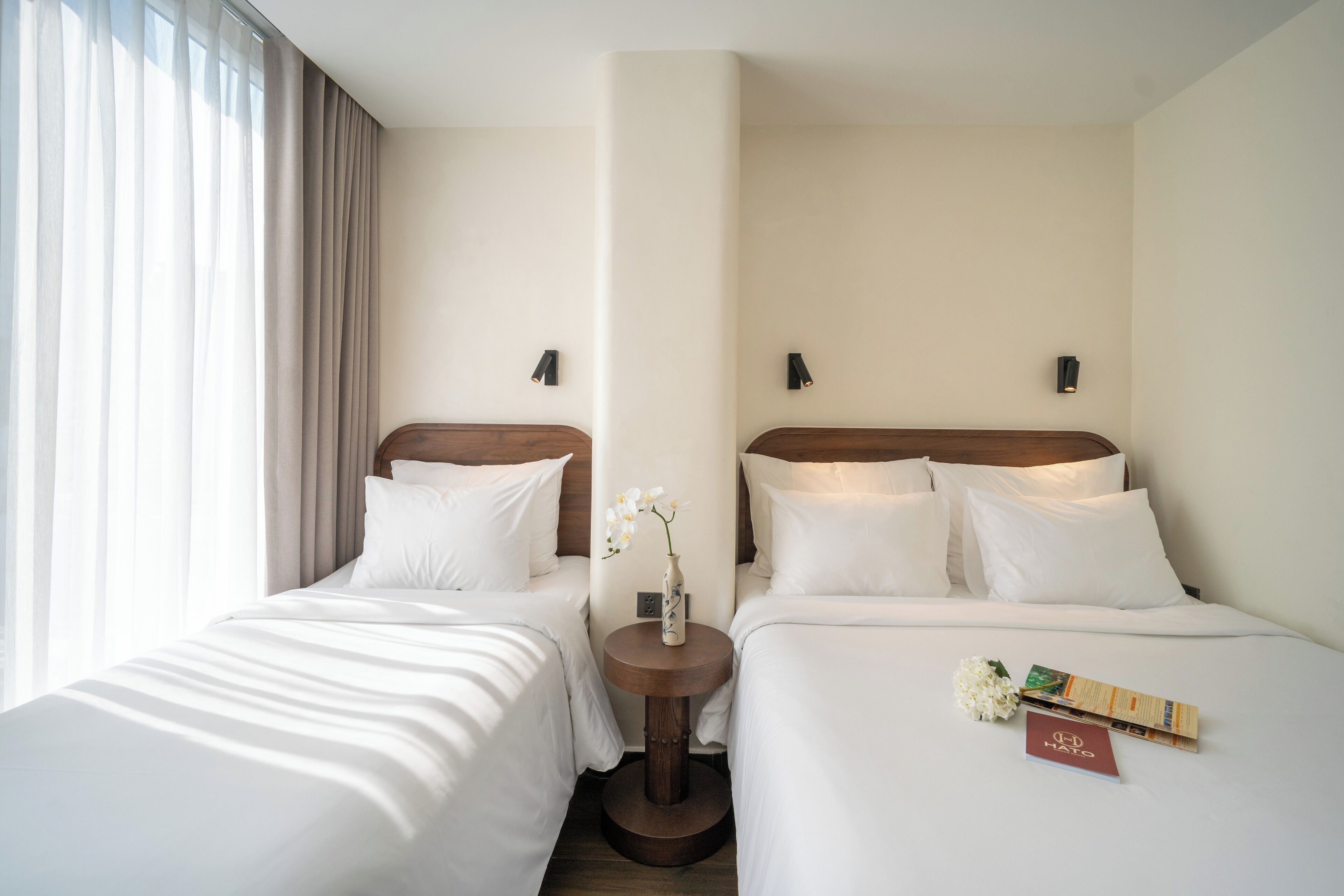 A hotel room with two beds, a round wooden table with a vase of flowers, and a small card on the bed.