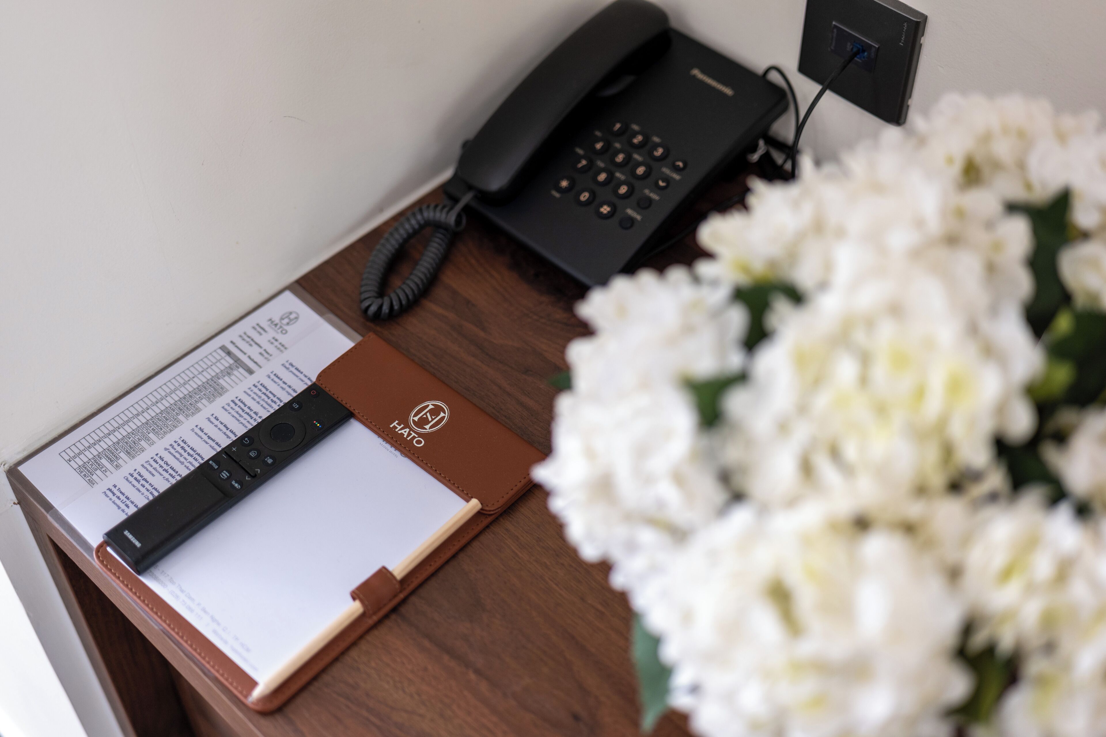 Un escritorio de madera con un teléfono negro, una libreta de cuero marrón y un ramo de flores blancas.