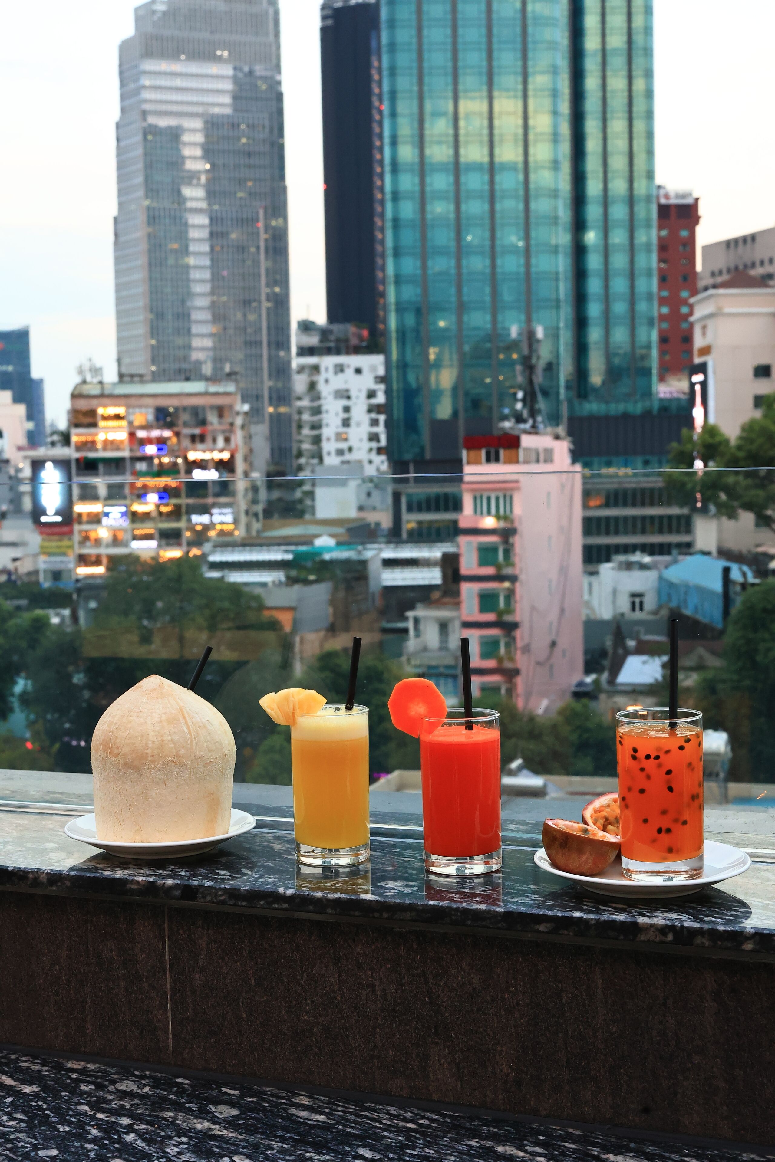 A cityscape view with a glass table displaying a coconut, two drinks, and a dessert.