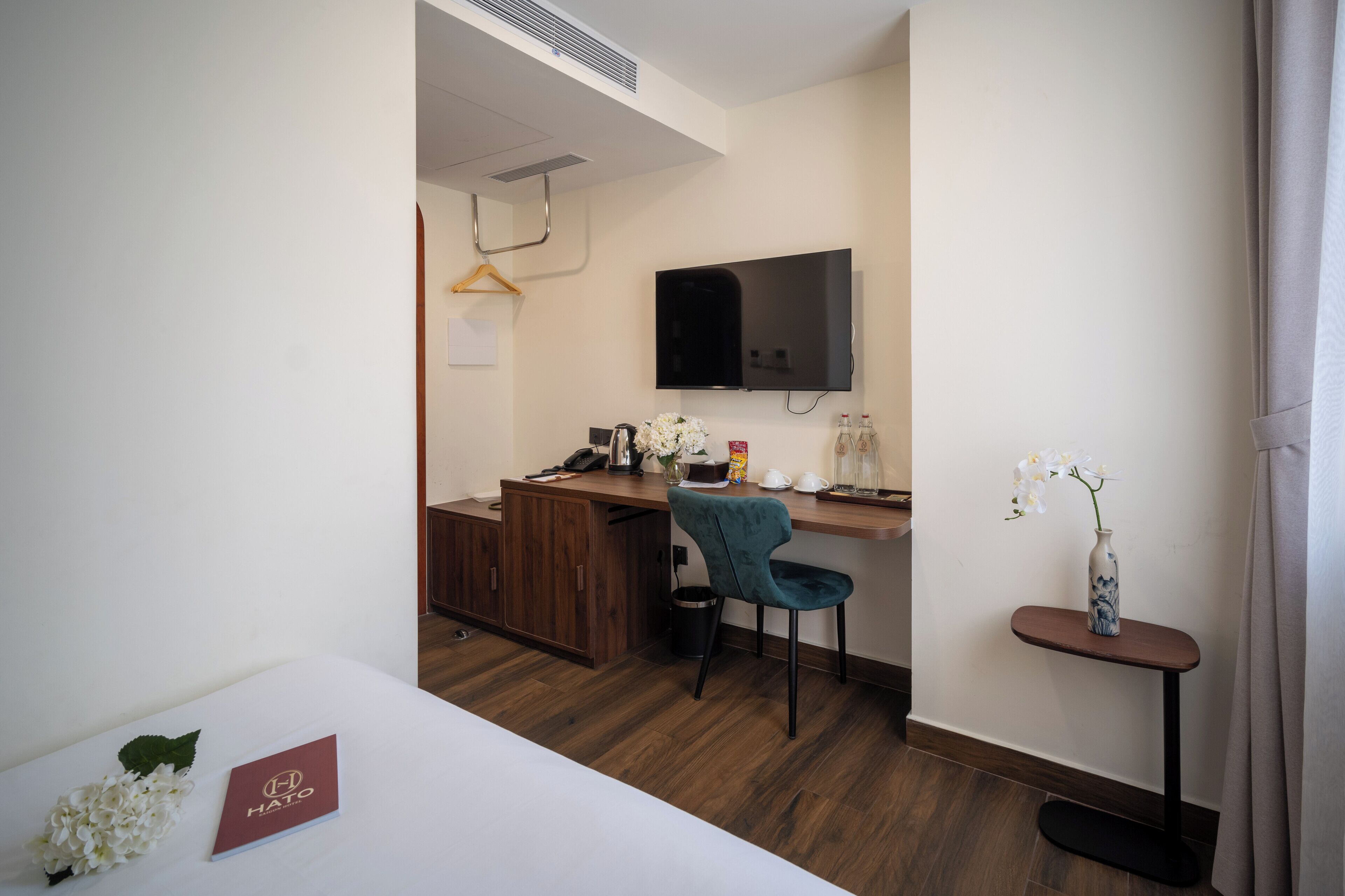 A hotel room with a wooden desk, a blue chair, a TV mounted on the wall, a vase with flowers, and a small table with a bottle.