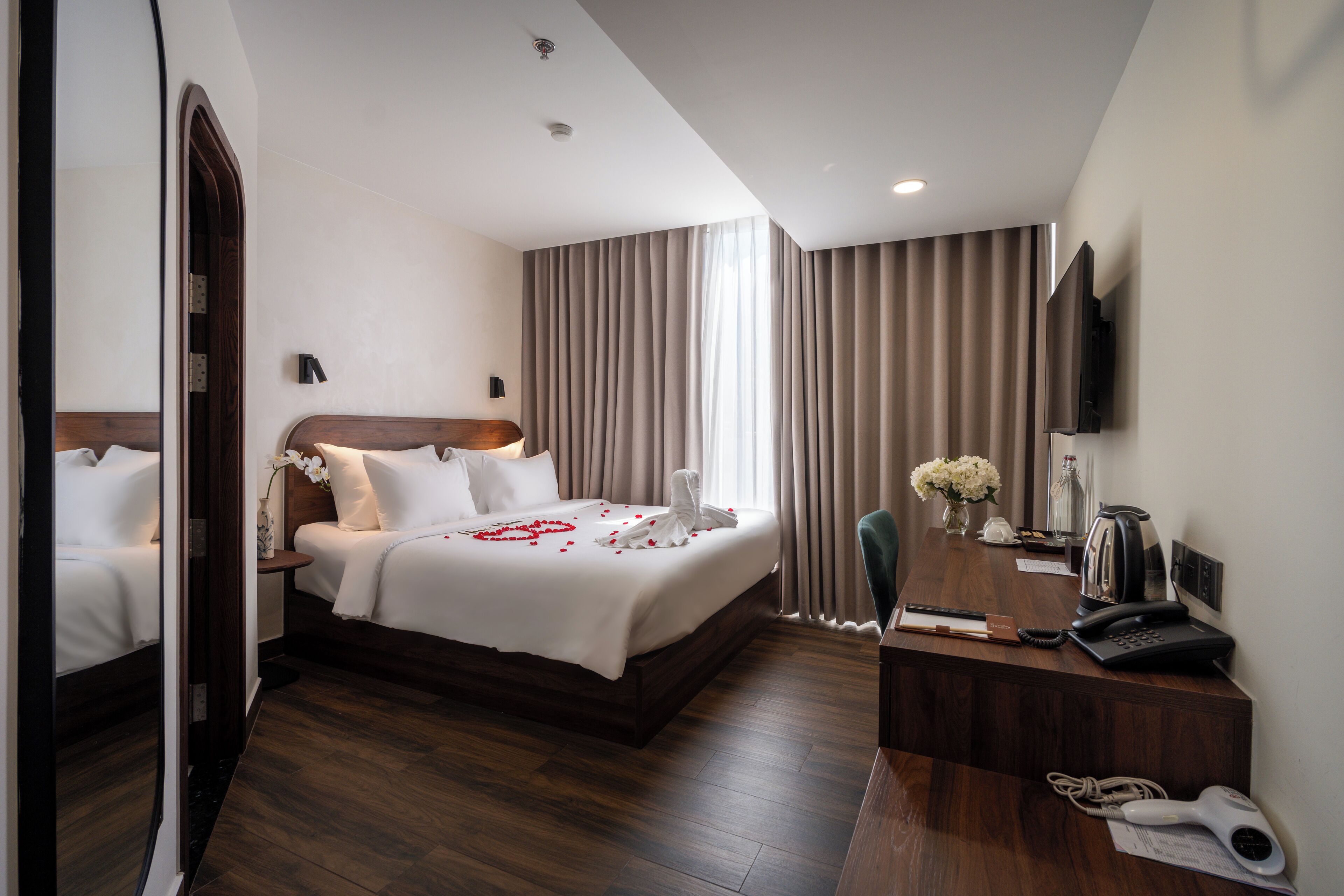 A hotel room with a large bed, a desk with a telephone and a coffee maker, a TV mounted on the wall, and a window with curtains.