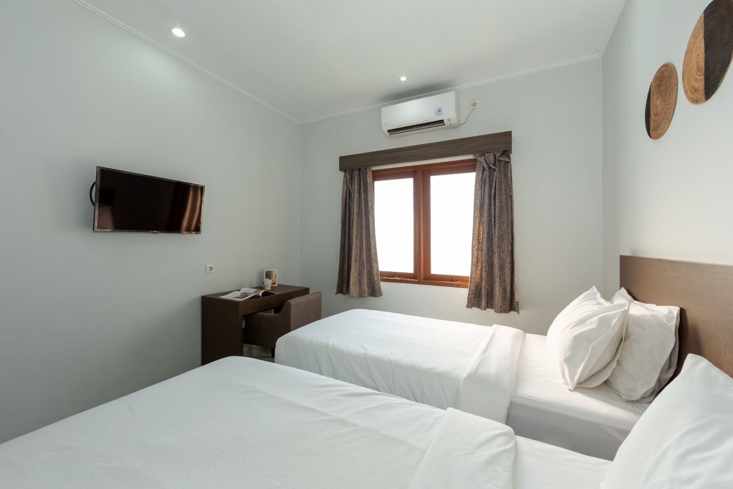A hotel room with two beds, a desk, a TV, an air conditioner, and a window with curtains.