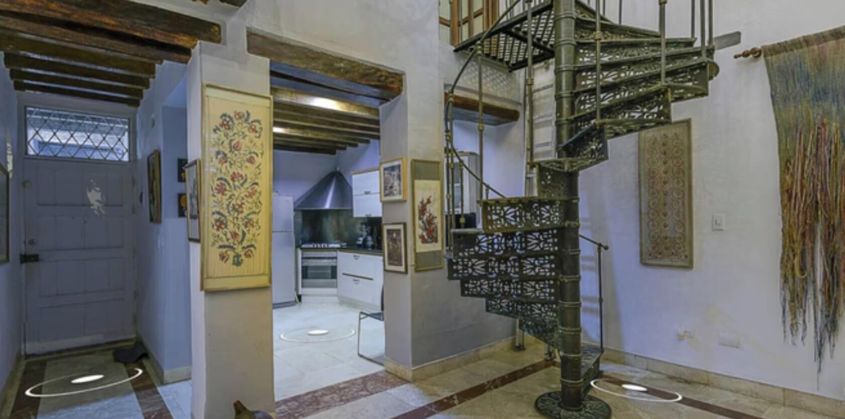 A spiral staircase in a hallway with a door, a kitchen in the background, and a patterned wall.