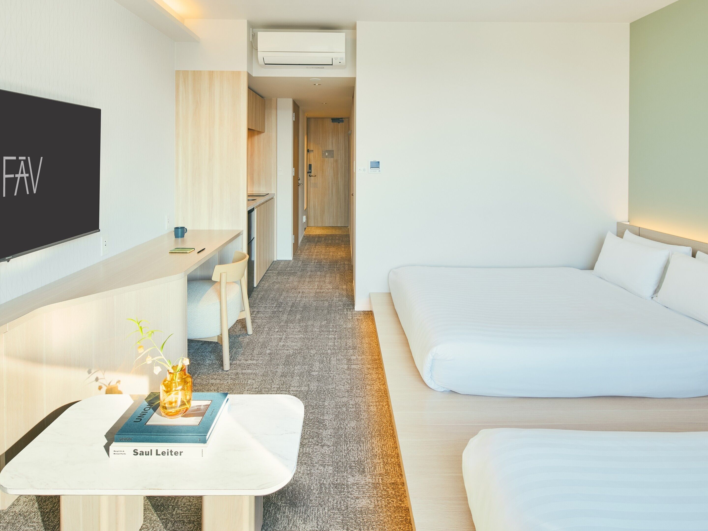 Una habitación de hotel con una cama, un escritorio con silla, un televisor y una mesita con un libro y un jarrón.