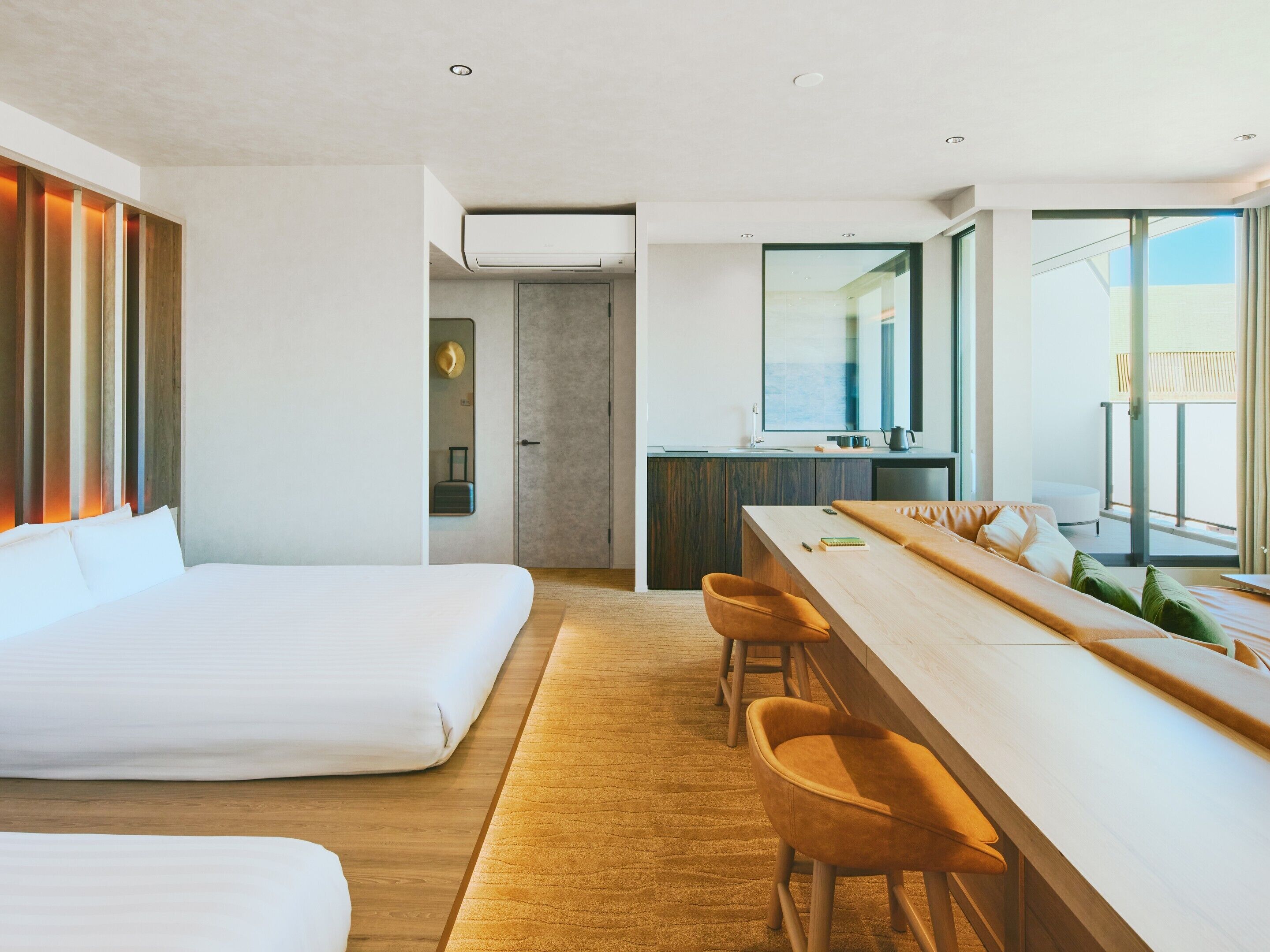 A modern hotel room with a large bed, a dining area with bar stools, a kitchenette, and a view of the outdoors.