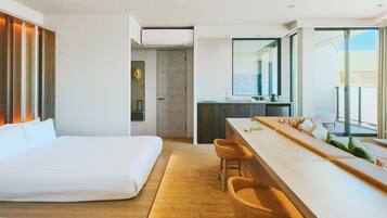 A modern hotel room with a large bed, a dining area with bar stools, a kitchenette, and a view of the outdoors.