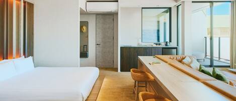 A modern hotel room with a large bed, a dining area with bar stools, a kitchenette, and a view of the outdoors.