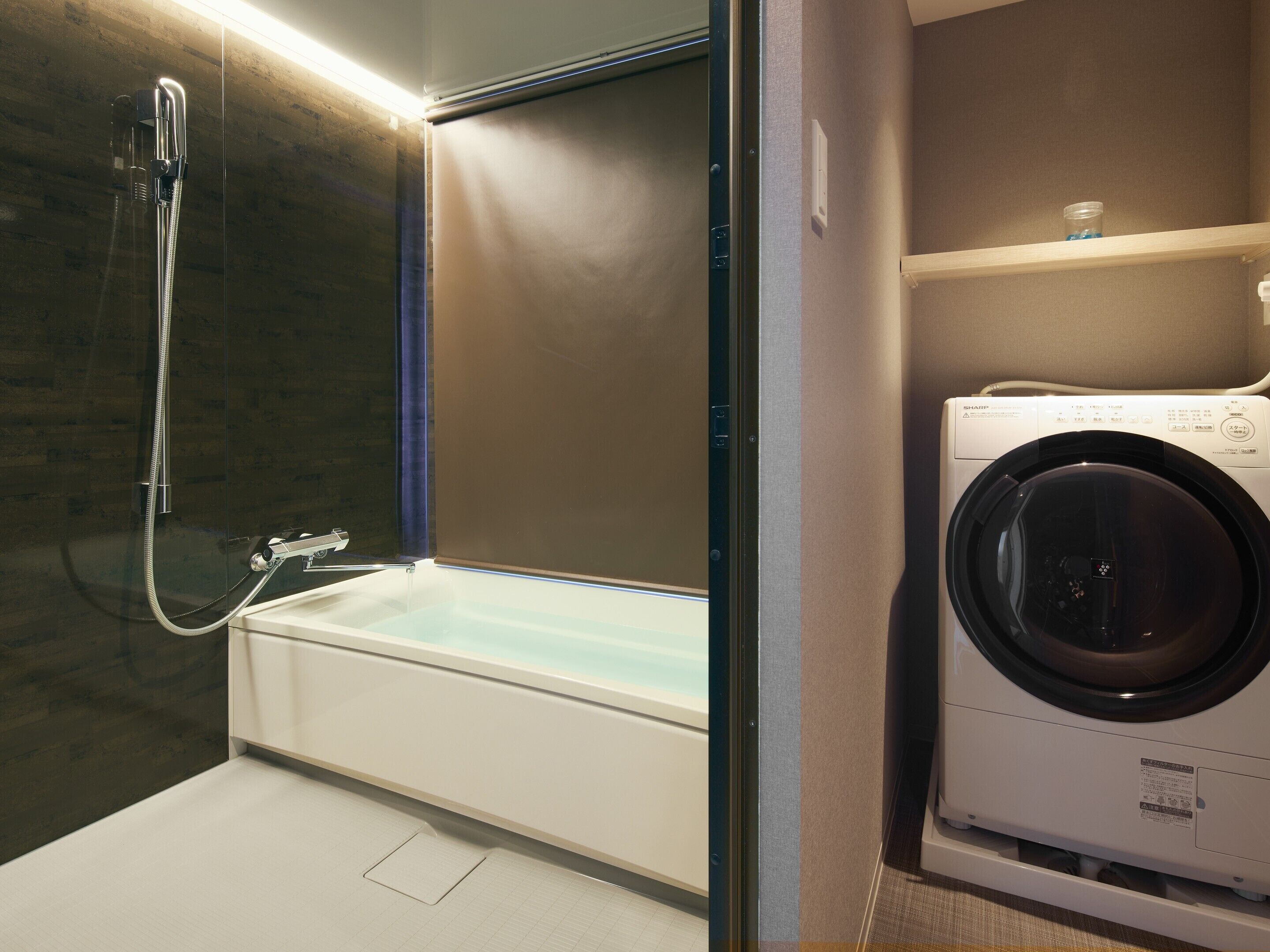 A compact bathroom with a glass-enclosed shower, a built-in washing machine, and a small shelf.
